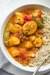 A bowl of seafood gumbo with shrimp, vegetables, and rice, garnished with herbs, next to a spoon on a white cloth.