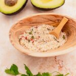 A wooden bowl filled with guacamole seasoning mix and a small wooden spoon. Halved avocados and cilantro leaves are visible in the background.