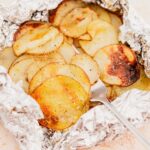 Sliced potatoes with seasoning are cooked and served in a foil packet, displayed with a fork.
