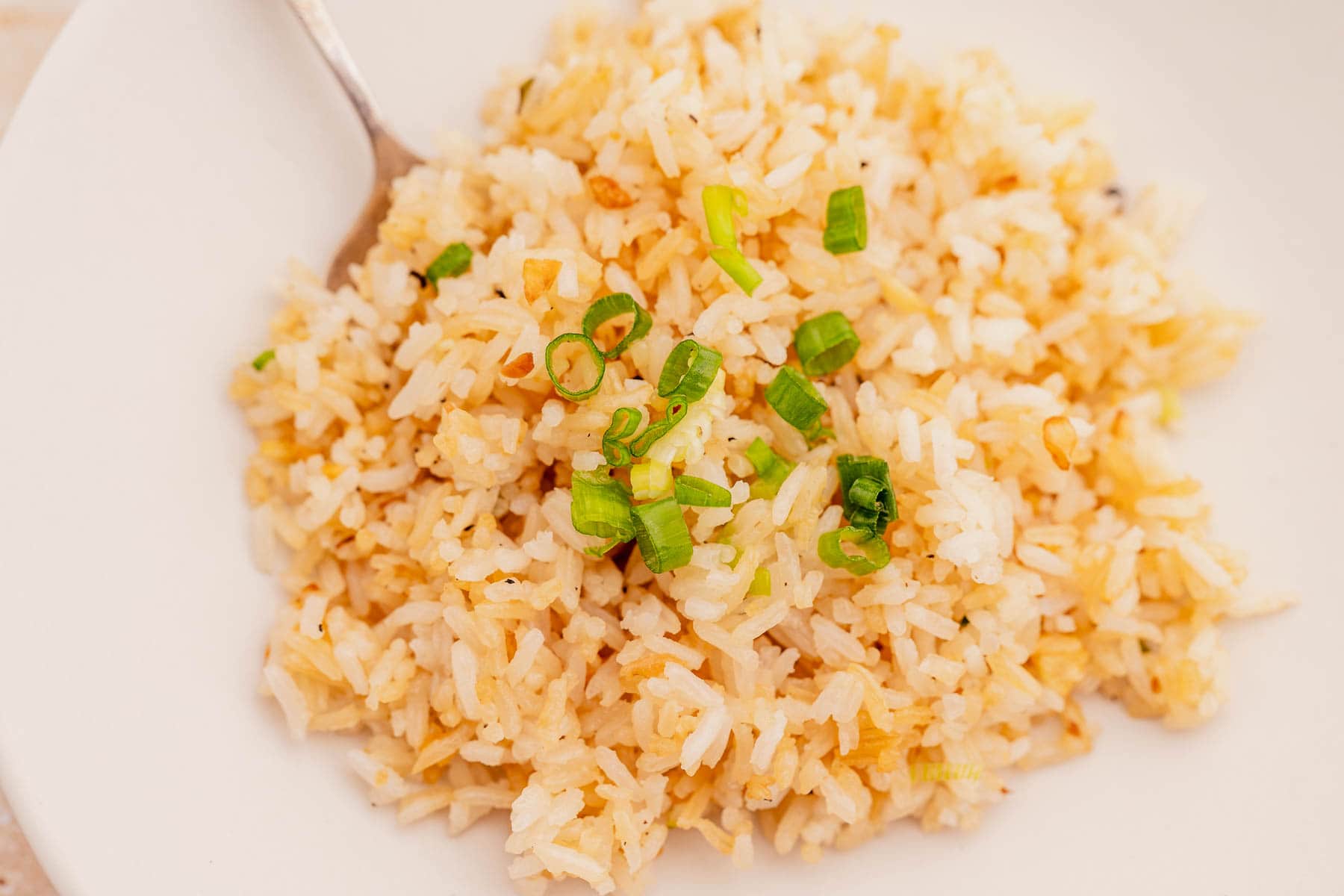 A plate of garlic fried rice garnished with chopped green onions, with a spoon on the side.