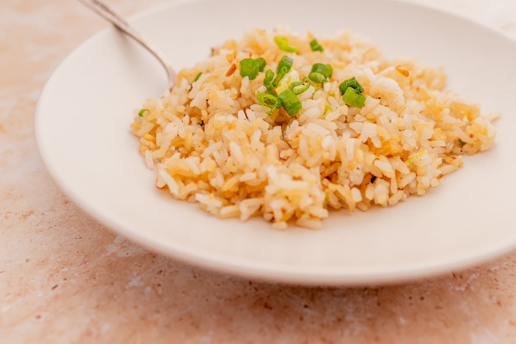 A white plate containing delicious garlic fried rice garnished with chopped green onions and accompanied by a spoon.