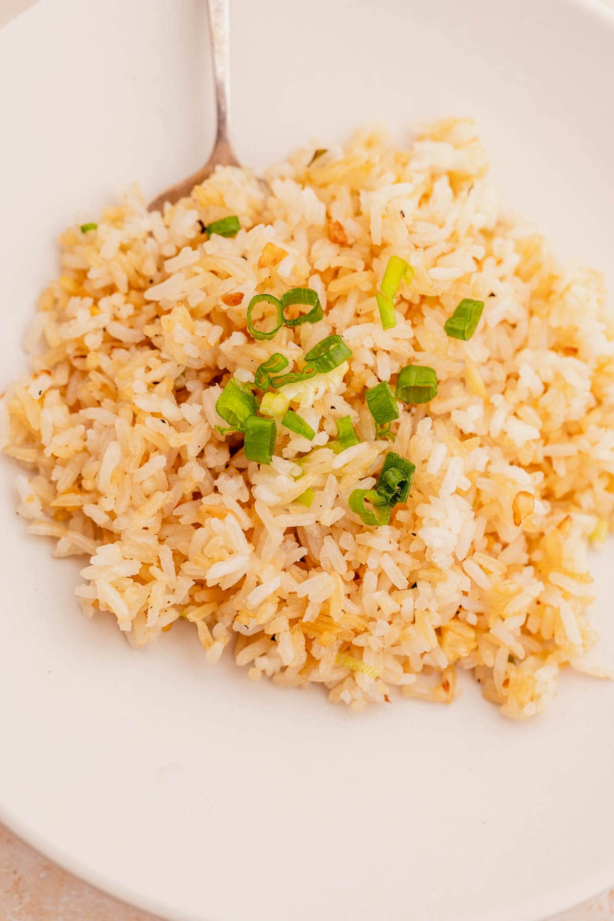 A white plate holds a serving of garlic fried rice, garnished with chopped green onions, with a spoon resting on the plate's edge.