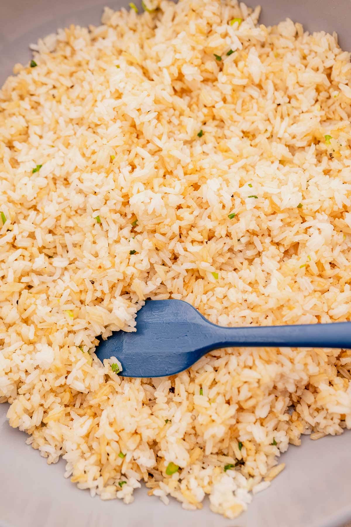 A bowl of garlic fried rice mixed with small green herbs, being stirred with a blue spatula.