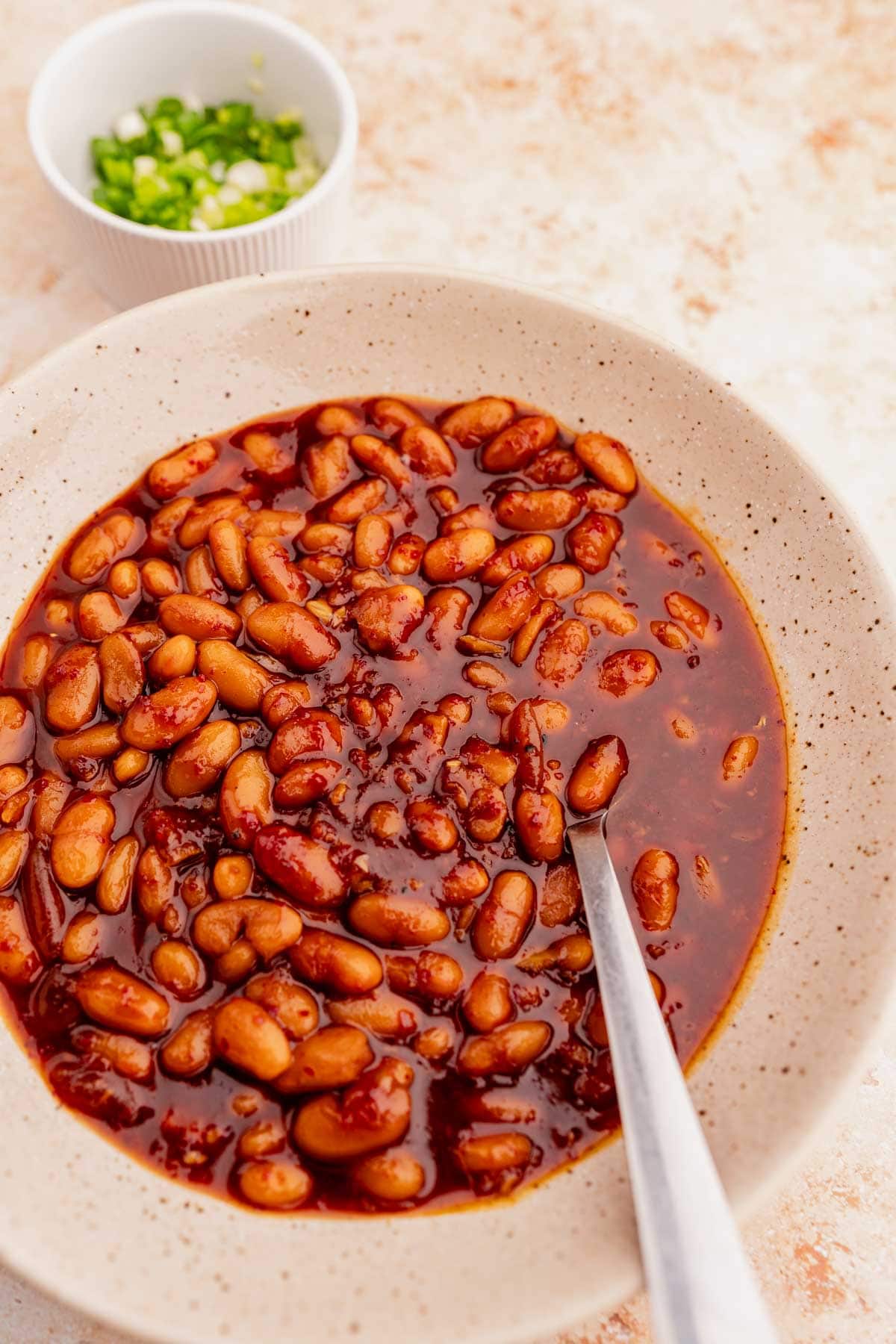 A ceramic bowl filled with Korean BBQ baked beans in a thick sauce, with a stainless steel spoon resting inside. A small dish of chopped green onions is in the background.