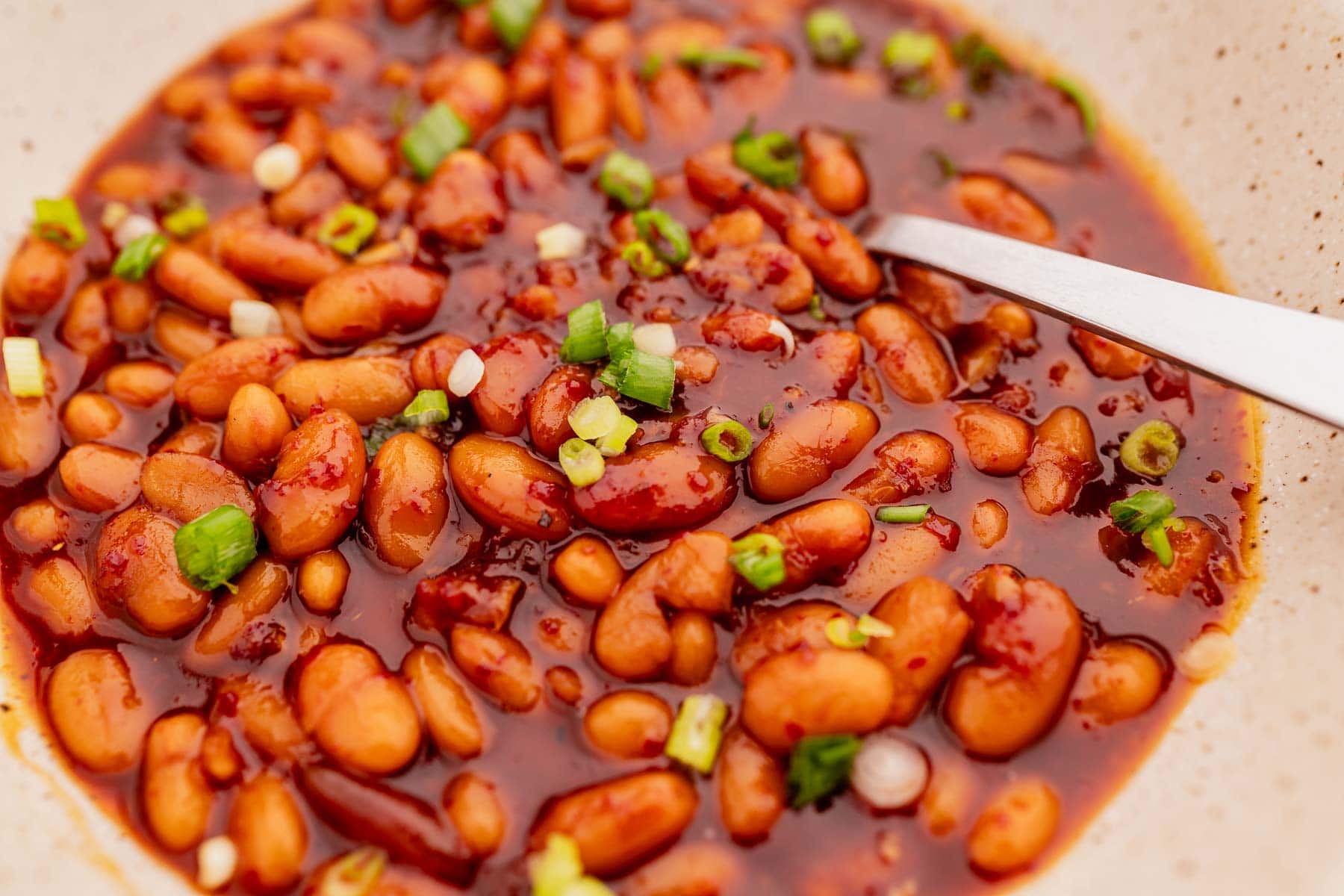 Close-up of a bowl of Korean BBQ baked beans in a thick sauce, garnished with chopped green onions, with a metal spoon resting in the bowl.