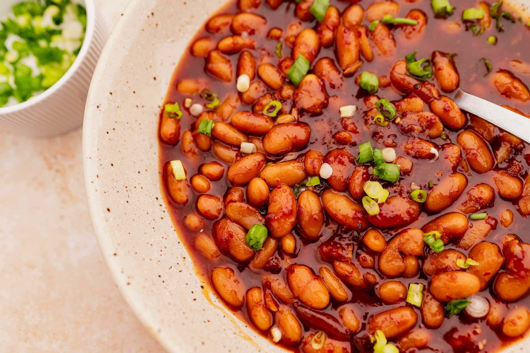 A bowl filled with Korean BBQ baked beans topped with chopped green onions, with a side dish of sliced green onions in the background.