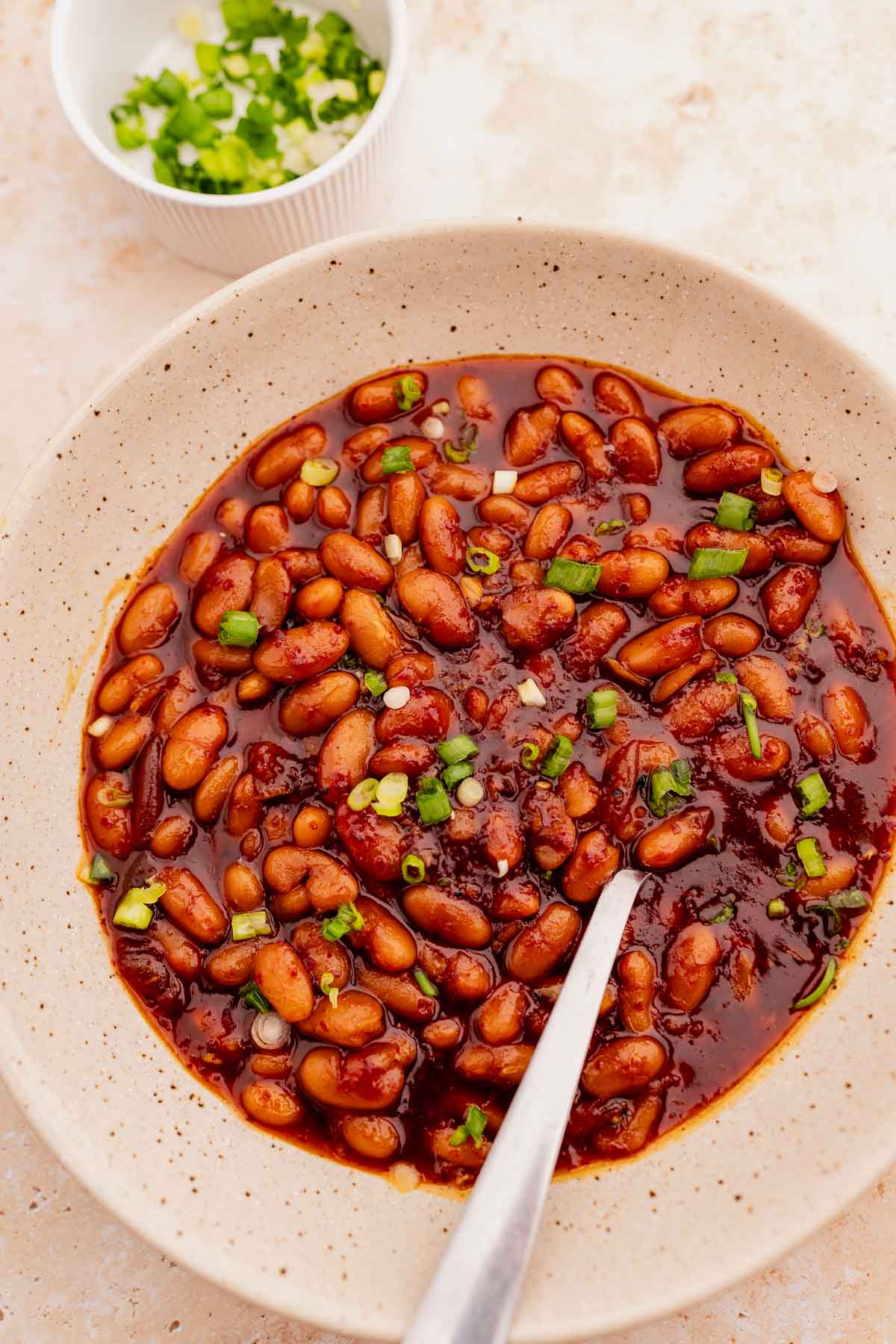 A bowl filled with Korean BBQ baked beans in a rich, red sauce, garnished with chopped green onions, with a spoon inside the bowl. A small bowl of additional chopped green onions is visible in the background.