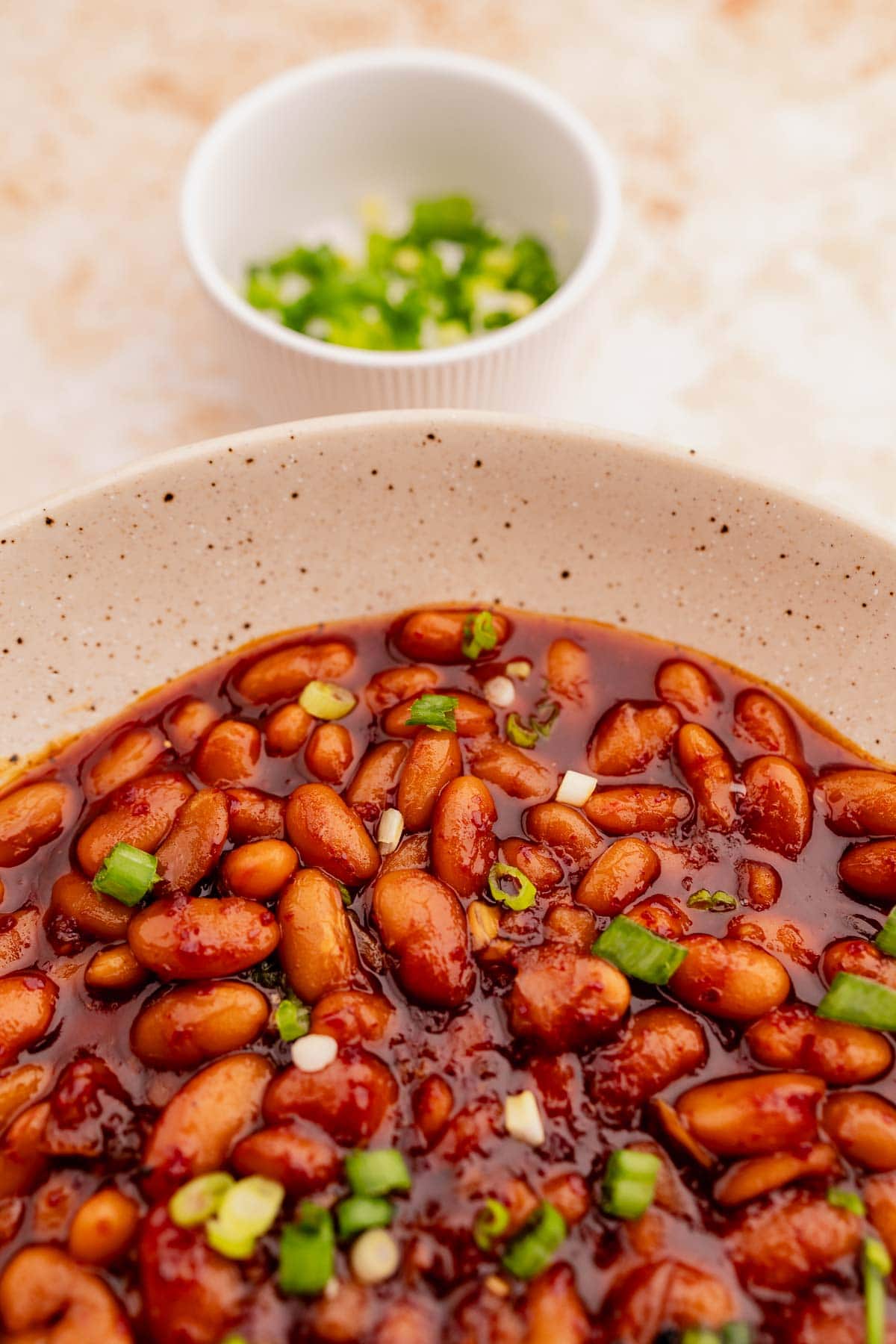 A bowl of Korean BBQ baked beans garnished with chopped green onions, with a small bowl of additional green onions in the background.