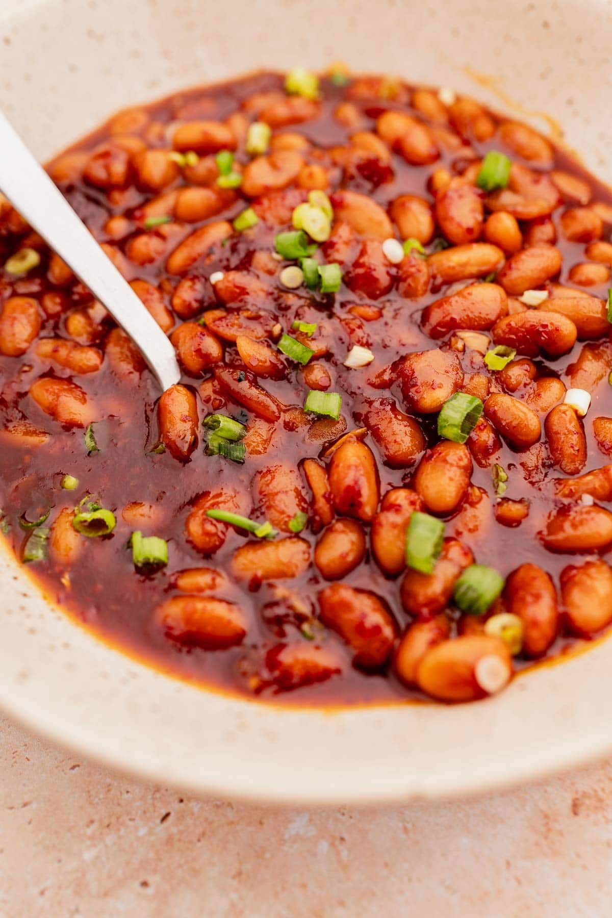 A bowl filled with Korean BBQ baked beans, garnished with chopped green onions, with a spoon resting on the side.