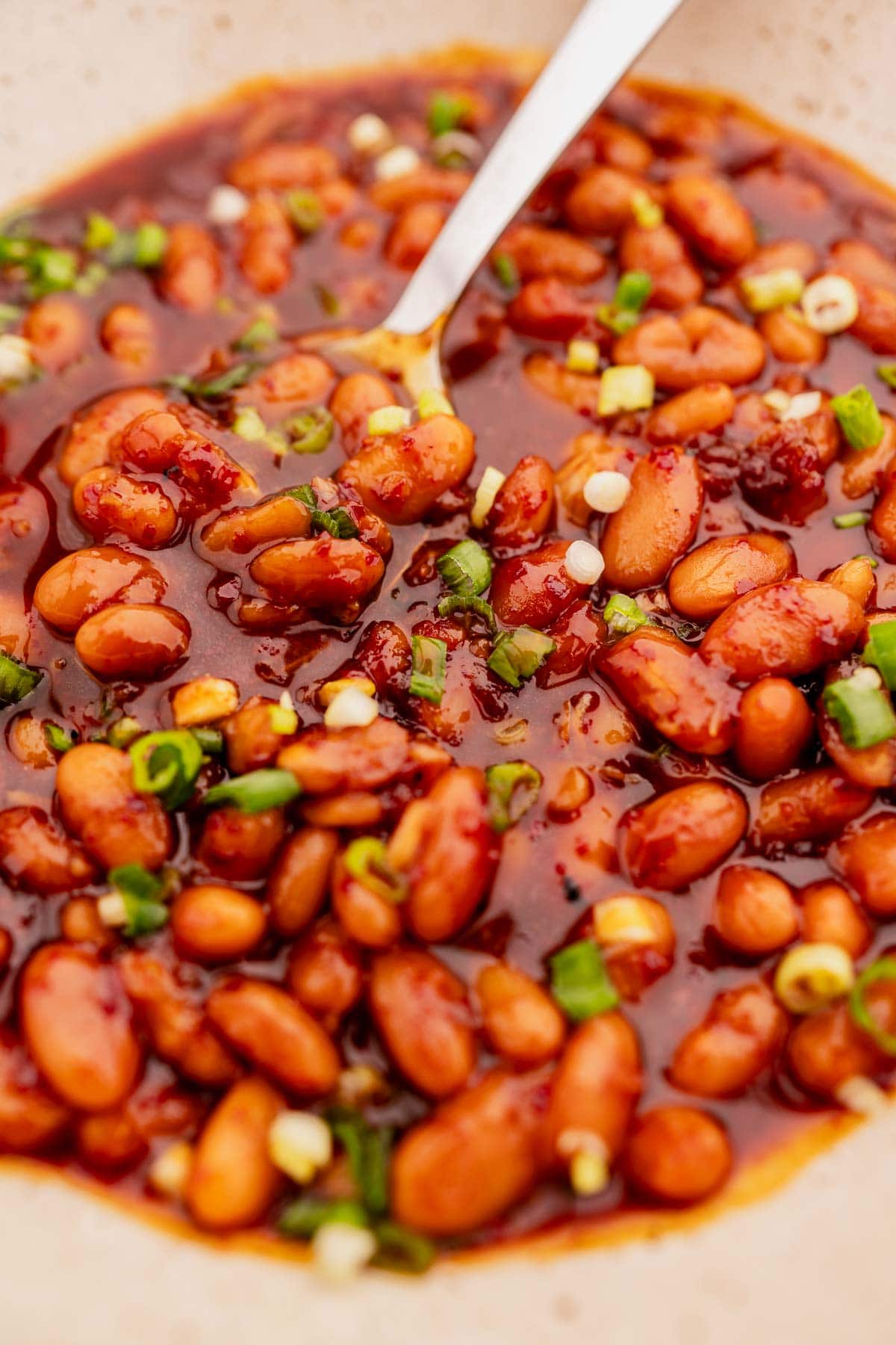 Close-up of a bowl of Korean BBQ baked beans in a thick sauce, garnished with chopped green onions. A silver spoon is partially submerged in the beans.