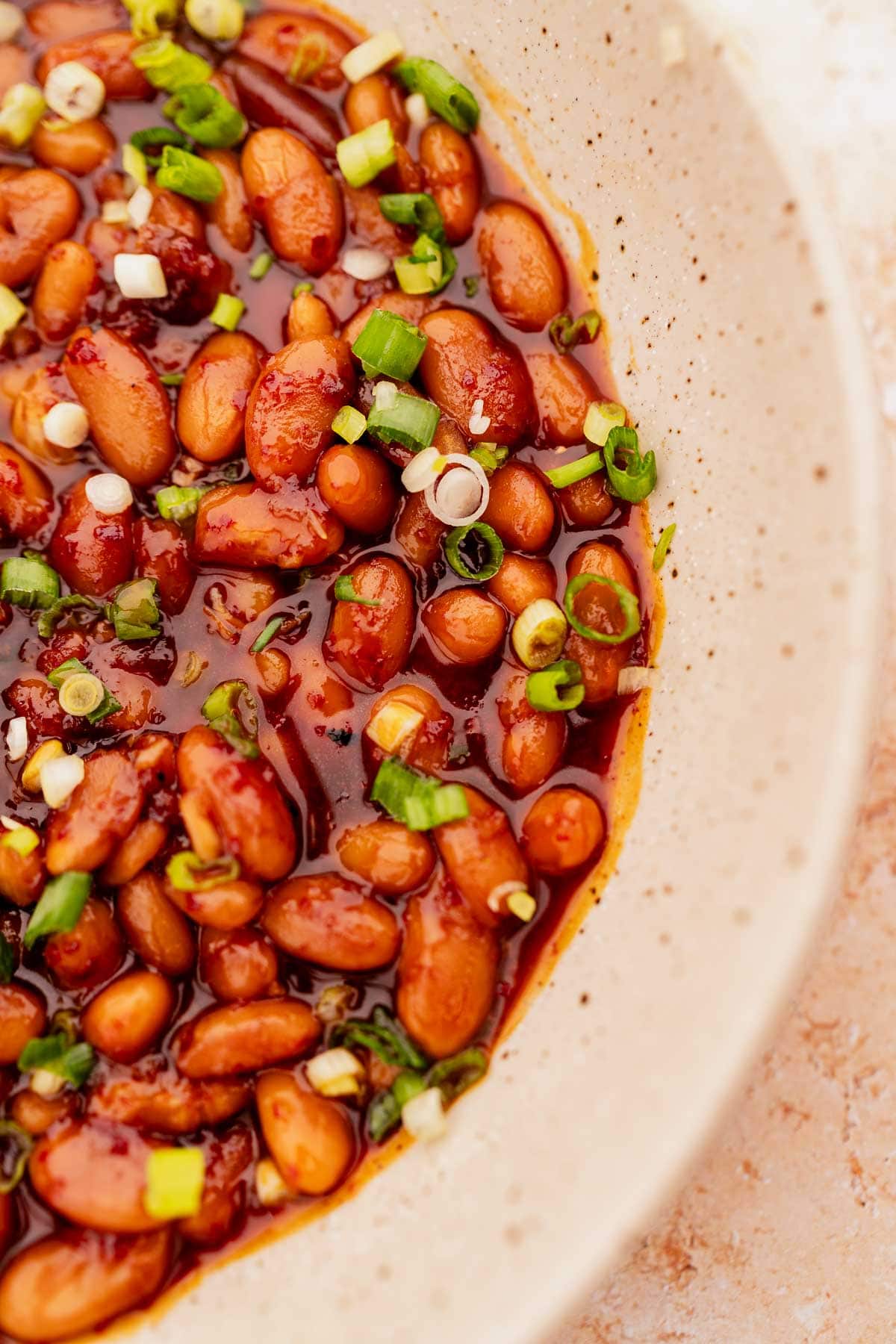 A close-up of Korean BBQ baked beans in a ceramic bowl, garnished with chopped green onions and mixed in a thick, reddish sauce.