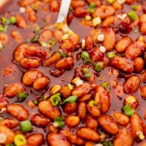 Close-up of Korean BBQ baked beans in a thick, reddish sauce garnished with chopped green onions and small pieces of chili, with a spoon partially submerged in the dish.