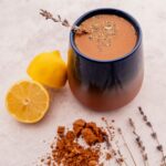 A blue and brown mug filled with frothy lavender hot chocolate garnished with delicate lavender sprigs. Next to the mug are a whole lemon, a lemon half, a pile of ground cinnamon, and pieces of dried lavender.