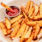 A plate of seasoned oven baked french fries served with a small metal cup of ketchup, with one crispy wedge dipped into the rich ketchup.