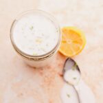 A jar filled with creamy coconut milk sauce, next to a sliced lemon and a spoon with sauce on it, sits on a light-colored surface. Lime zest flecks are visible in the dressing.