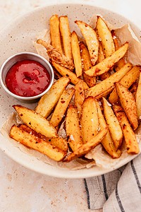 A bowl of seasoned oven baked french fries on parchment paper, served with a small metal cup of ketchup on the side.