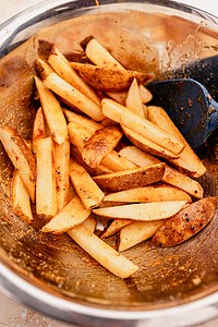 Potato wedges coated with spices in a large metal mixing bowl, resembling oven baked french fries, with a blue spatula partially visible.