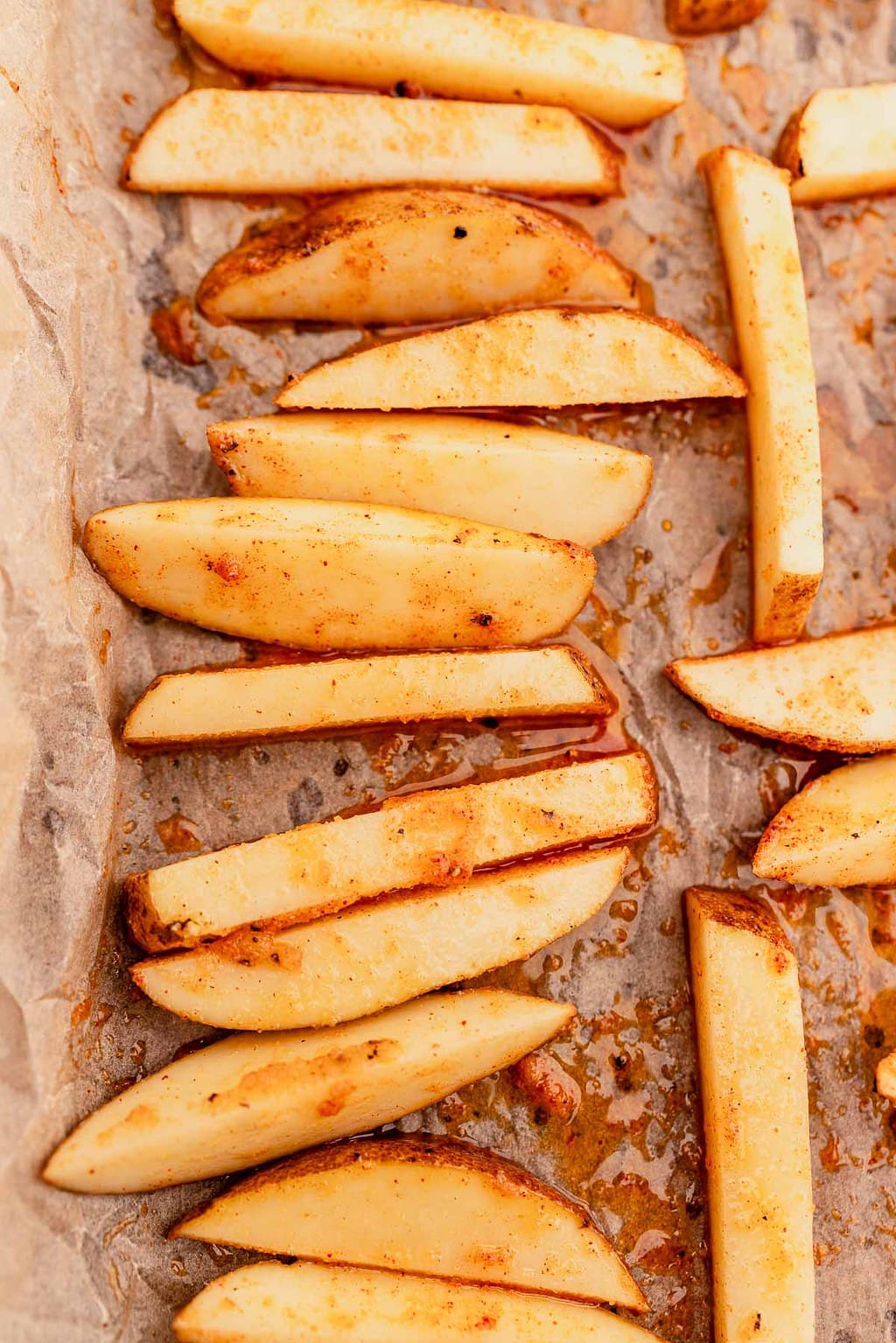 Potato wedges coated in seasoning are arranged on a sheet of parchment paper, ready for baking into crispy oven baked french fries.
