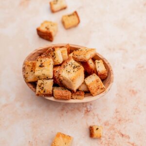 A small wooden bowl filled with seasoned, gluten-free croutons on a light, speckled surface, with a few croutons scattered around.