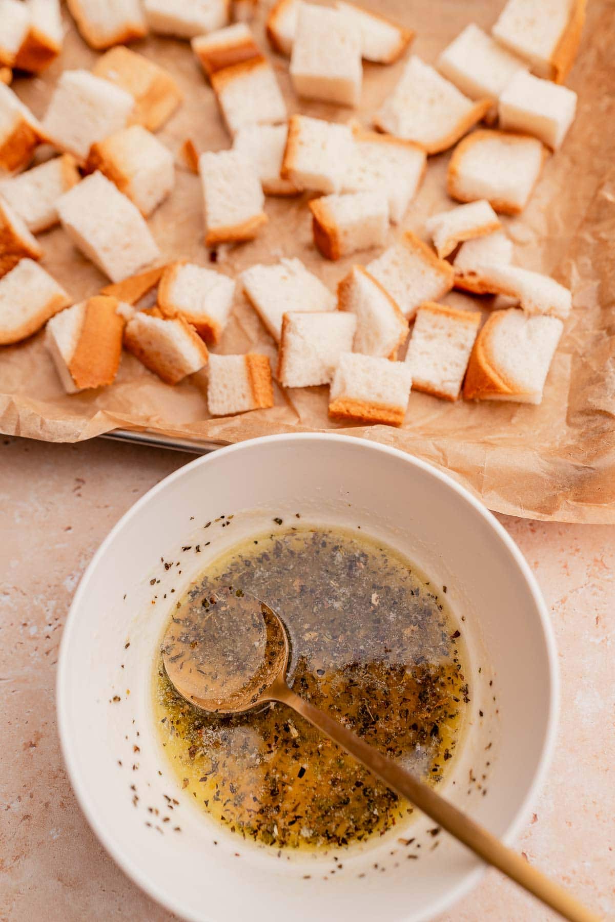 A bowl with an herb and oil mixture next to a baking tray lined with parchment paper and cubed gluten-free croutons.