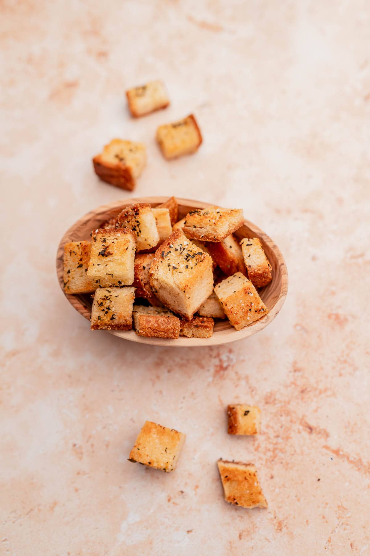 A wooden bowl filled with seasoned, gluten-free croutons is placed on a light-colored, textured surface. Some croutons are scattered around the bowl.