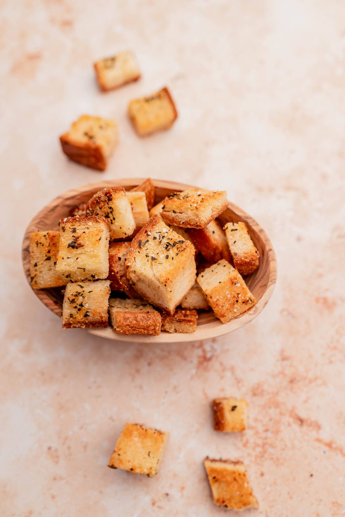 A wooden bowl filled with seasoned, gluten-free croutons, with some croutons scattered around the bowl on a light-colored surface.