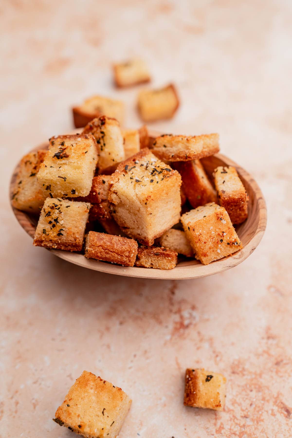 A wooden bowl filled with seasoned, gluten-free croutons, with a few croutons scattered on a beige surface around it.