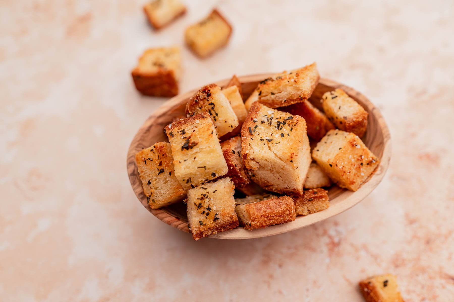 A wooden bowl filled with seasoned gluten-free croutons on a beige surface, with some croutons scattered around.