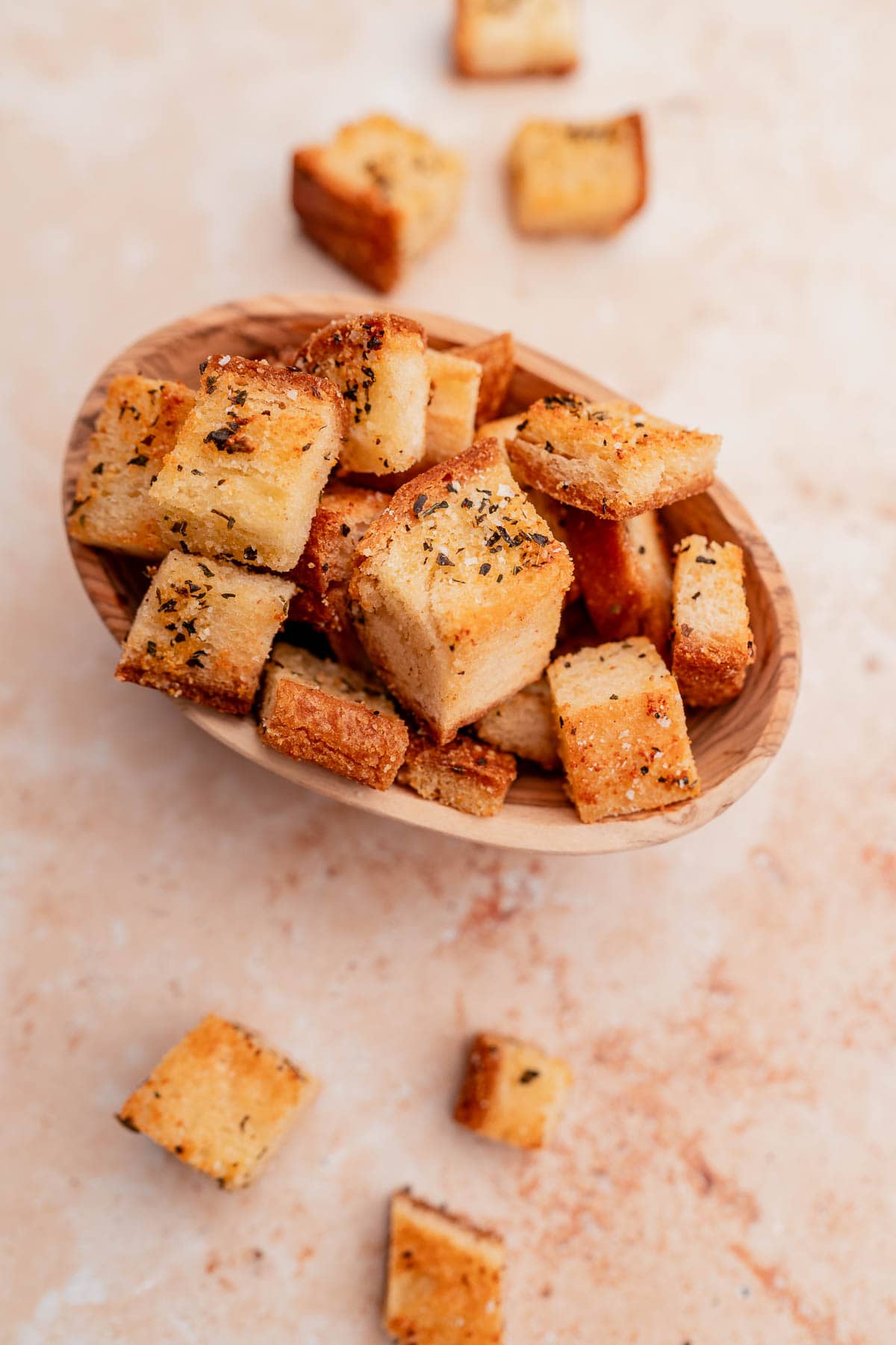 A wooden bowl filled with seasoned gluten-free croutons. Some croutons are scattered on the light-colored surface.