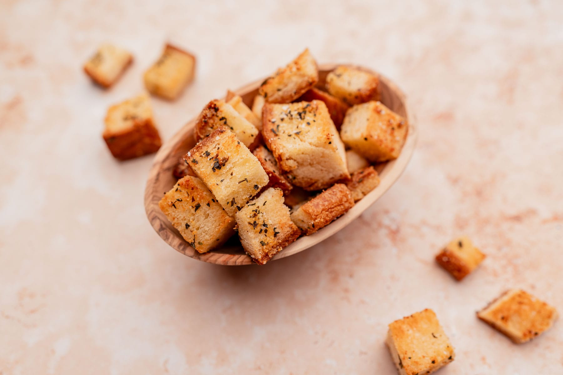 A wooden bowl filled with seasoned gluten free croutons, placed on a light beige surface, with some croutons scattered around the bowl.