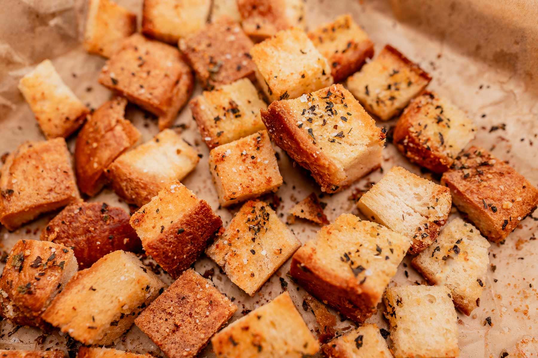 Close-up of seasoned gluten-free bread cubes on a baking sheet, likely croutons. The cubes are golden brown with visible herbs and spices.