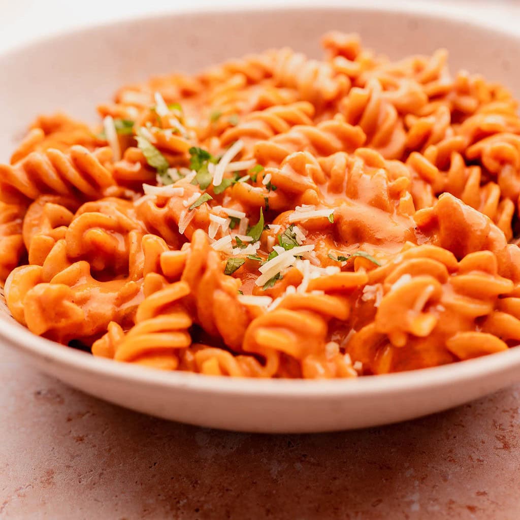 A close-up of a bowl of sun-dried tomato pasta rotini covered in marinara sauce and topped with grated cheese and herbs.