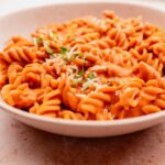 A close-up of a bowl of sun-dried tomato pasta rotini covered in marinara sauce and topped with grated cheese and herbs.