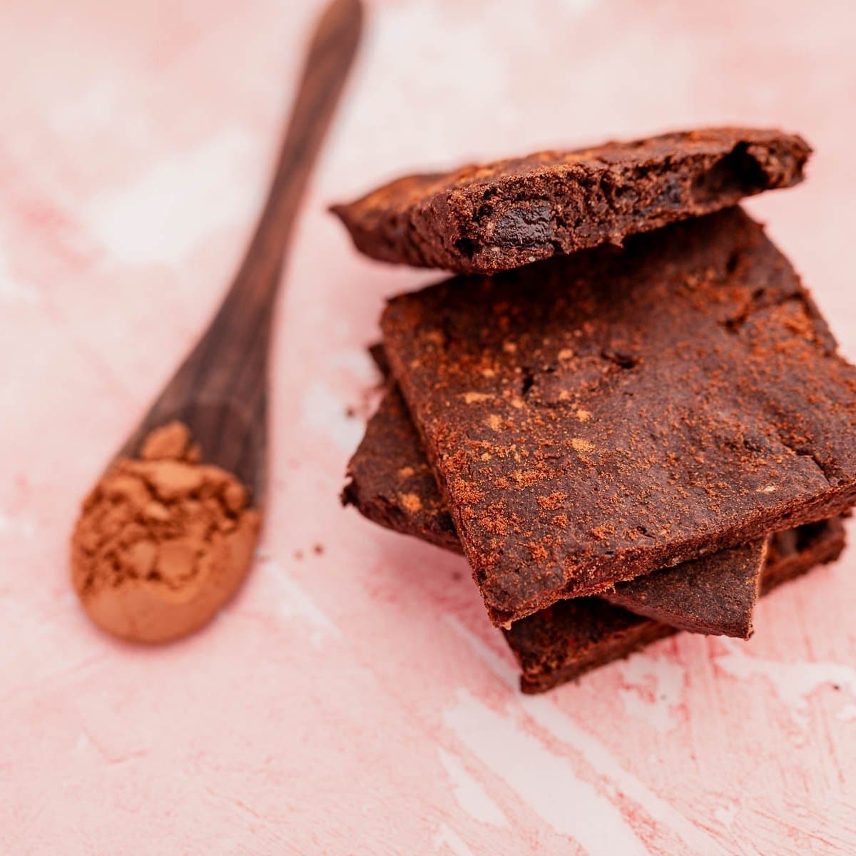 Stack of three chocolate brownies next to a wooden spoon filled with cocoa powder on a pink textured surface.