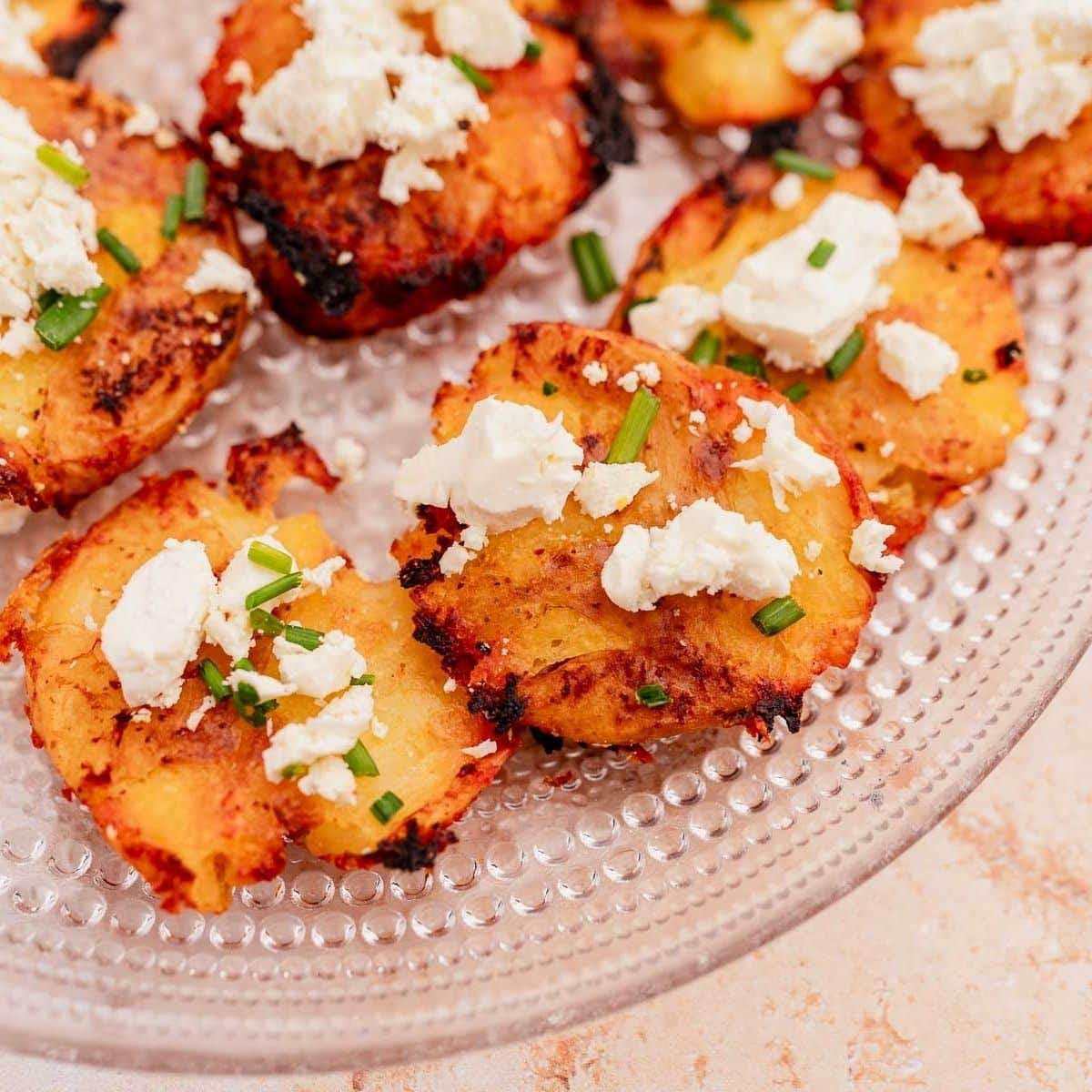 Crispy smashed potatoes topped with white crumbled cheese, chopped green herbs, and a drizzle of hot sauce on a clear textured plate.