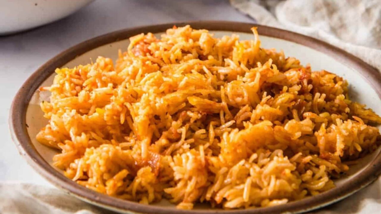 A close-up of a pot with cooked jollof rice with a person's hand holding a red spatula.