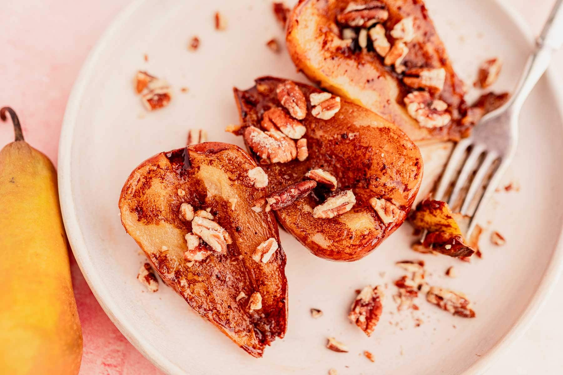 Grilled chocolate balsamic glazed pear halves topped with chopped pecans rest on a white plate, accompanied by a fork and a whole pear nearby.