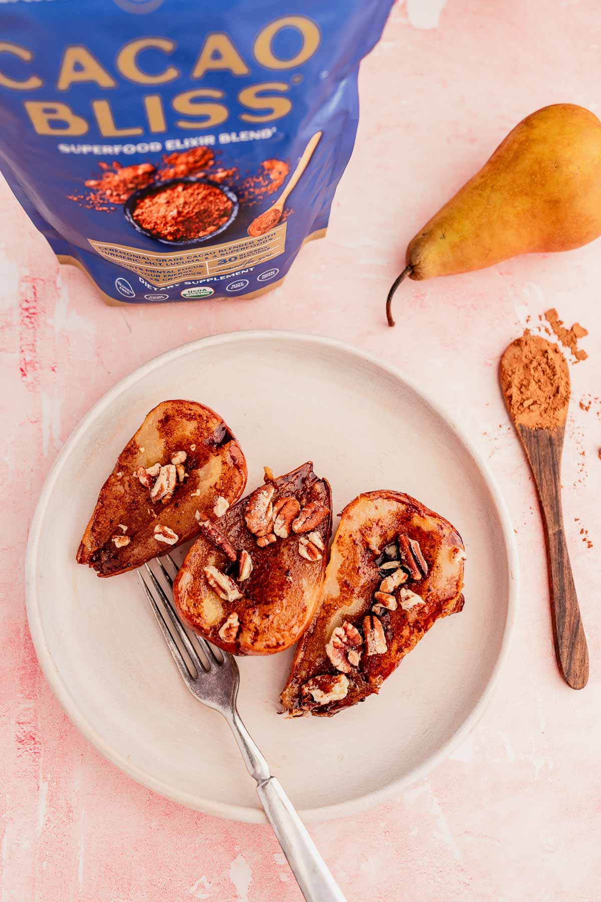 Plate of roasted pear halves topped with pecans and cocoa powder, accompanied by a fork; chocolate balsamic glazed pears add a rich twist. A pouch of Cacao Bliss and a fresh pear are in the background.