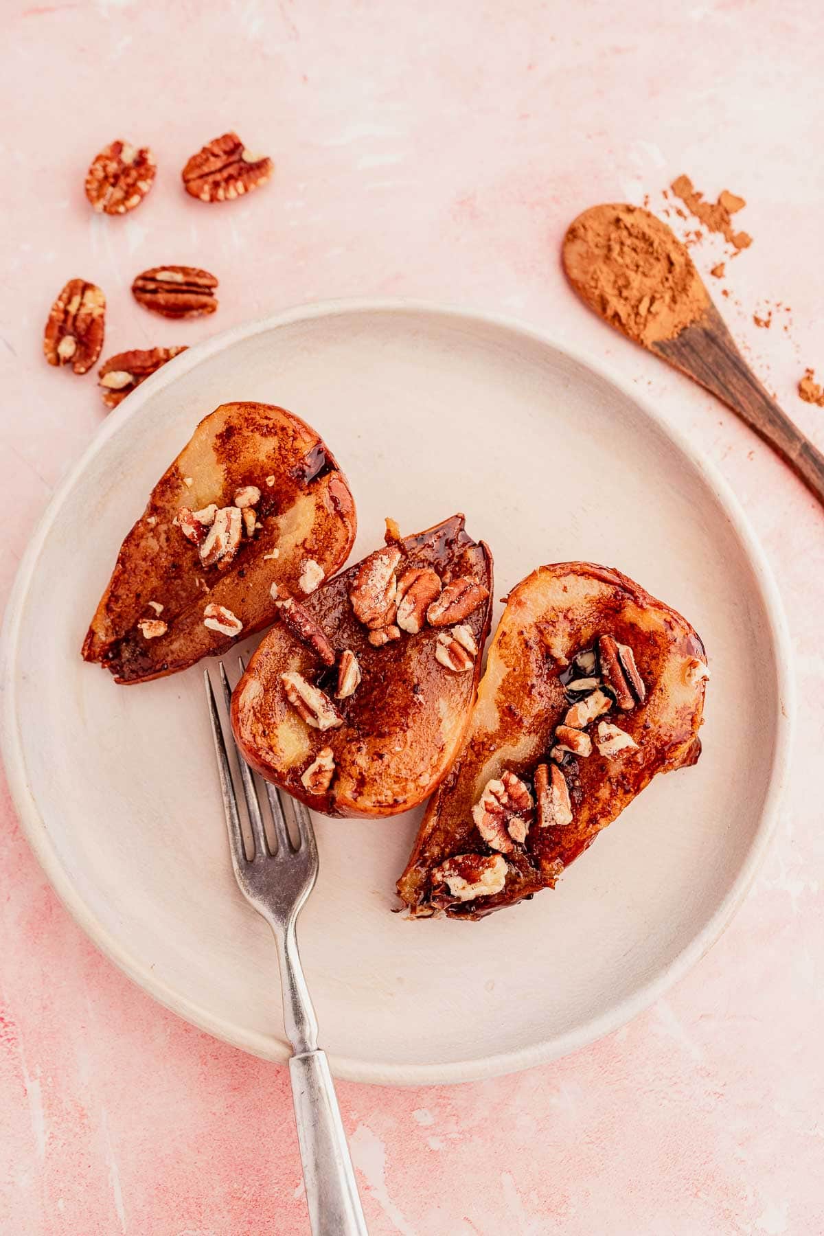 Three pear halves baked with cinnamon and pecans, drizzled with a chocolate balsamic glaze, rest elegantly on a white plate. Nearby, a fork, pecans, and a wooden spoon dusted with cinnamon are artfully arranged on the pink surface.