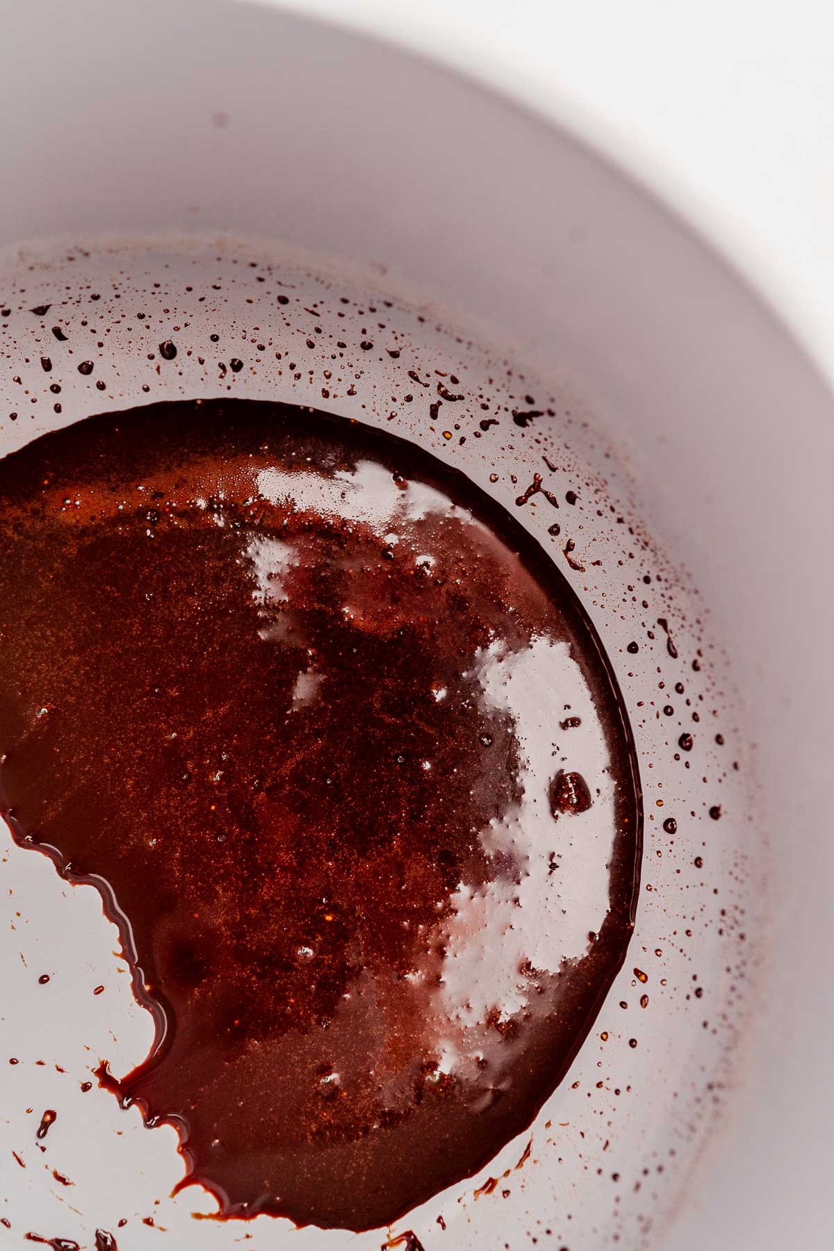 Close-up of a bowl with partially melted reddish-brown chocolate, showing specks and a glossy texture on the surface, reminiscent of the rich glaze found on chocolate balsamic glazed pears.