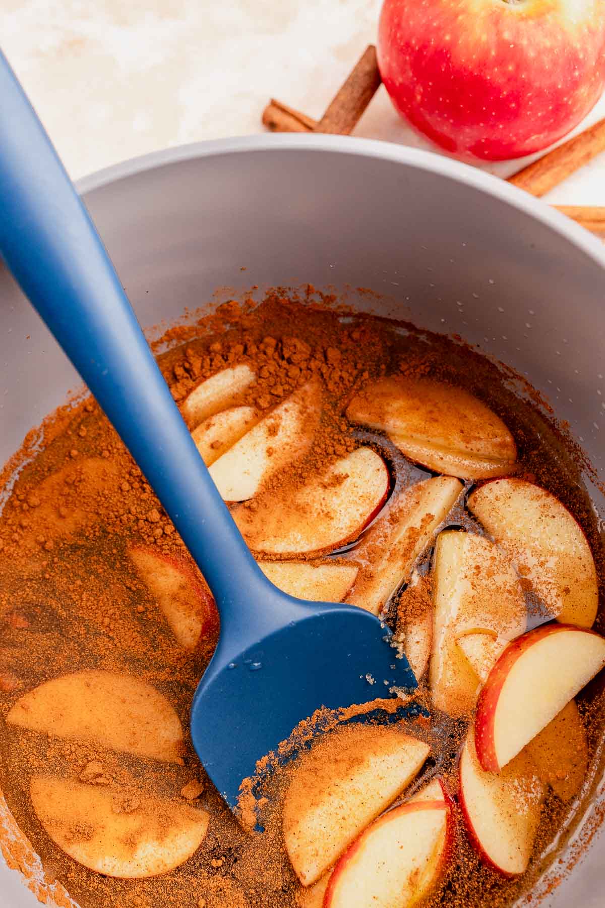 Sliced apples with cinnamon in a pan are stirred with a blue spatula, enhanced by a hint of Starbucks apple crisp syrup. An apple and cinnamon sticks are visible in the background.