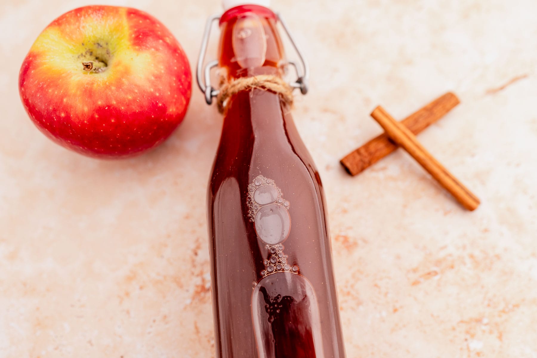 A glass bottle of dark liquid, reminiscent of Starbucks apple crisp syrup, lies next to a red apple and two cinnamon sticks on a light surface.