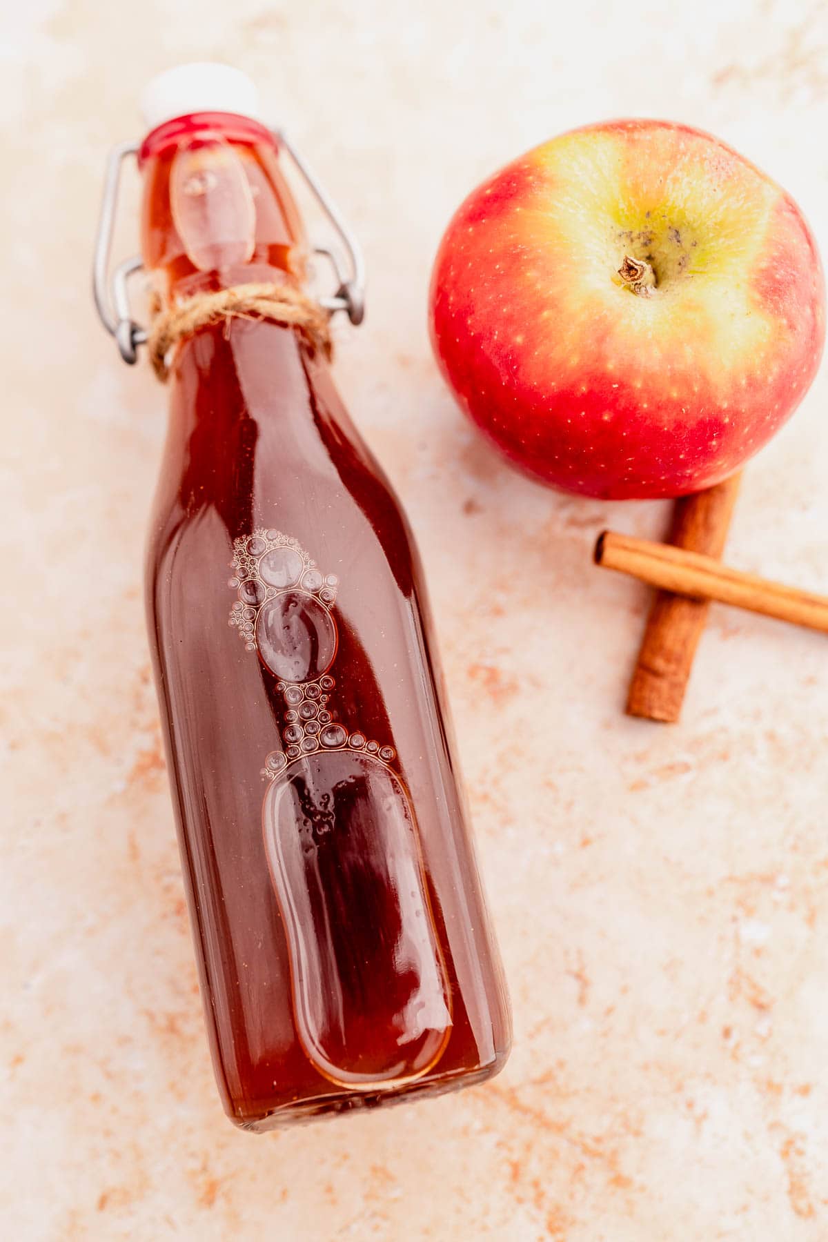 A glass bottle with a swing top, filled with Starbucks apple crisp syrup, sits beside a red apple and two cinnamon sticks on a light, textured surface.