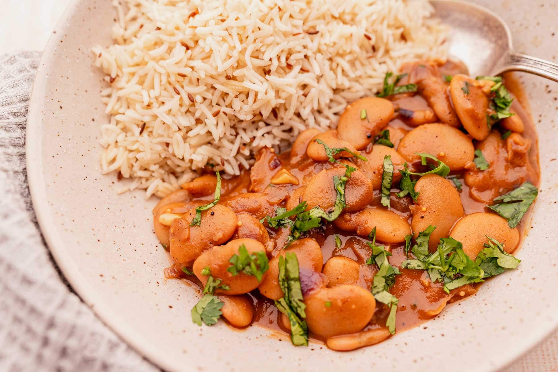 Butter bean curry and white rice rest in a tan ceramic bowl with a large silver spoon.