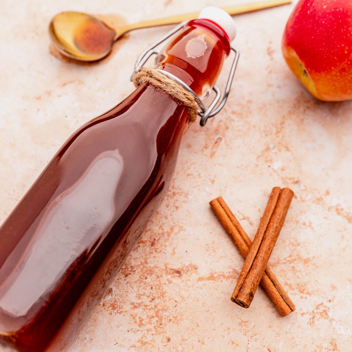 A glass bottle of dark liquid, a spoon with residue, a red apple, and two cinnamon sticks on a light surface.