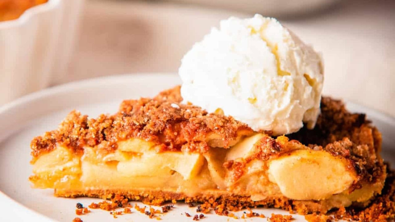 A close-up photo of a sliced apple pie with graham cracker crust on a white plate, topped with a scoop of vanilla ice cream. 