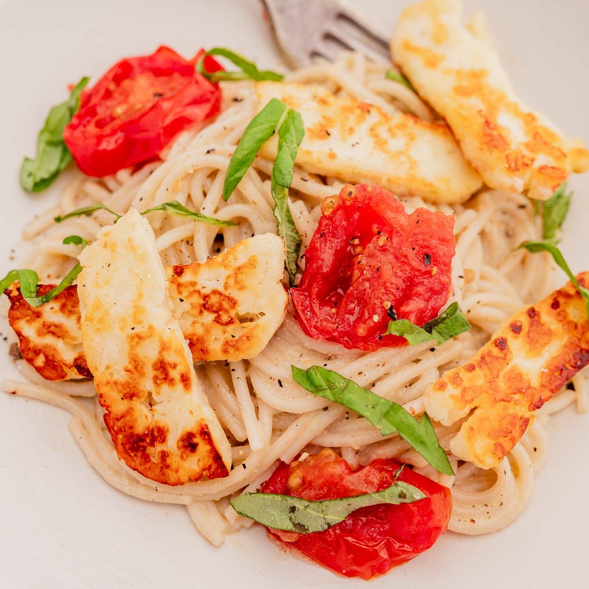 A plate of spaghetti topped with grilled halloumi, chopped tomatoes, and basil, with a fork in the background.