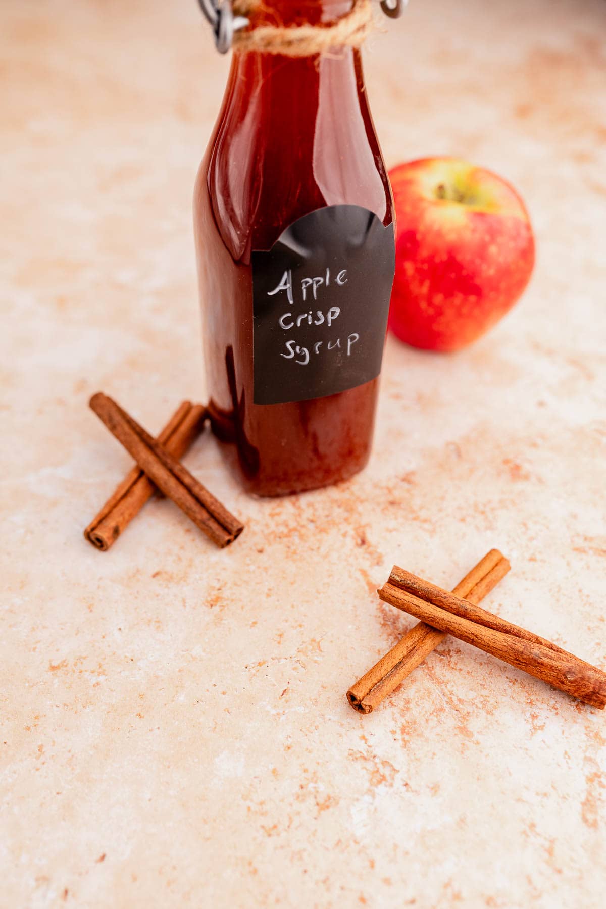 A bottle labeled "Starbucks Apple Crisp Syrup" with two cinnamon sticks is placed next to a red apple on a textured surface.