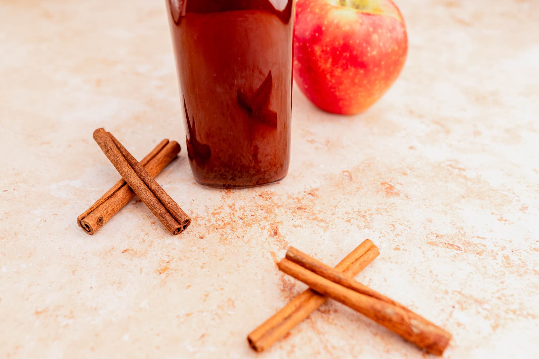 A glass bottle of dark liquid, flanked by an apple and paired cinnamon sticks, rests elegantly on a light marble surface. It's a scene reminiscent of the rich flavors captured in Starbucks' delightful apple crisp syrup.