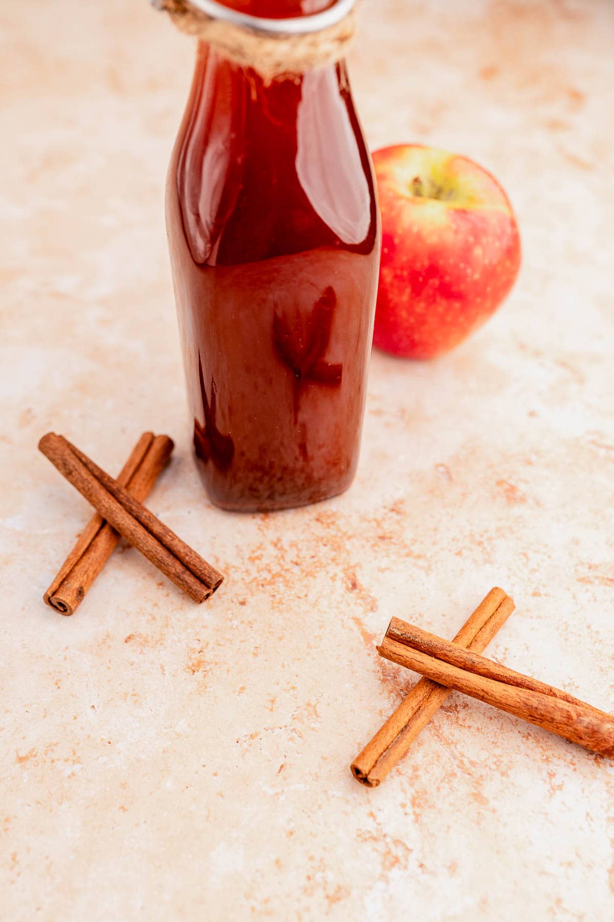 A glass bottle of brown liquid, reminiscent of Starbucks apple crisp syrup, sits next to a red apple and two crossed pairs of cinnamon sticks on a light surface.